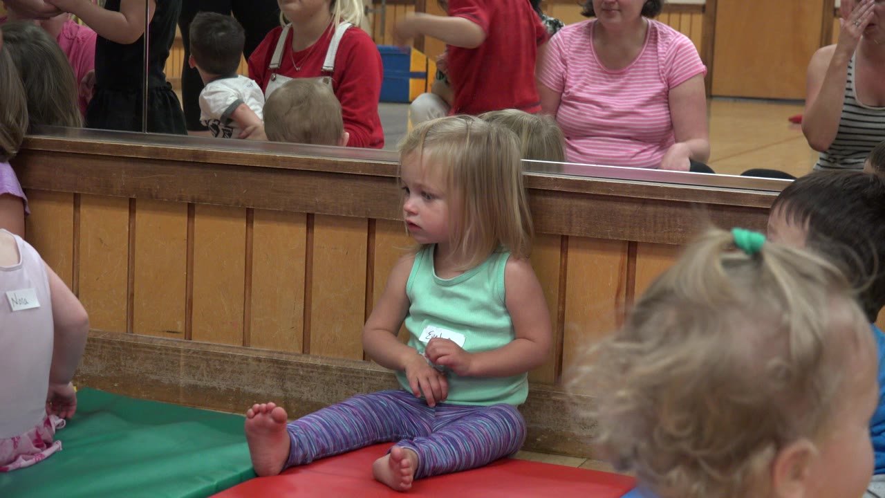 Toddler girl sitting on colored play mats near other children.