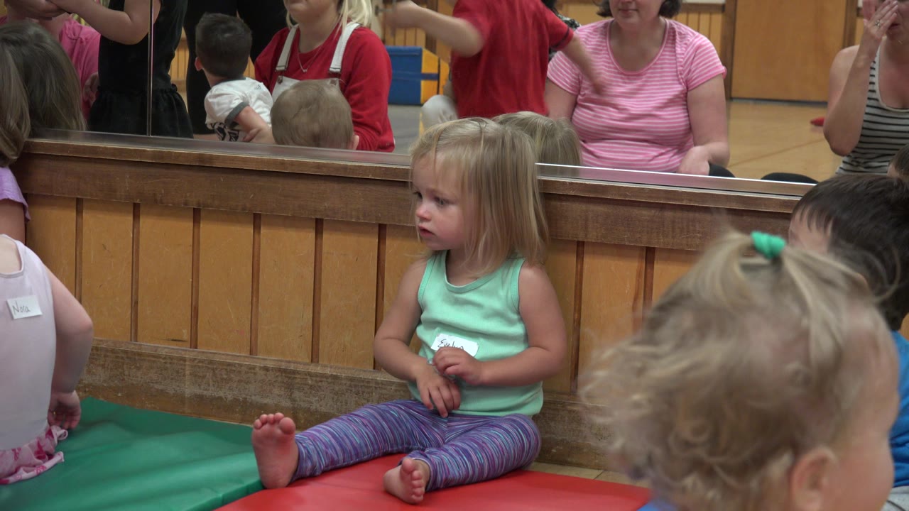 The seated girl remains centered on the mats as a child's head fills the foreground and adults are reflected in the mirror behind her.