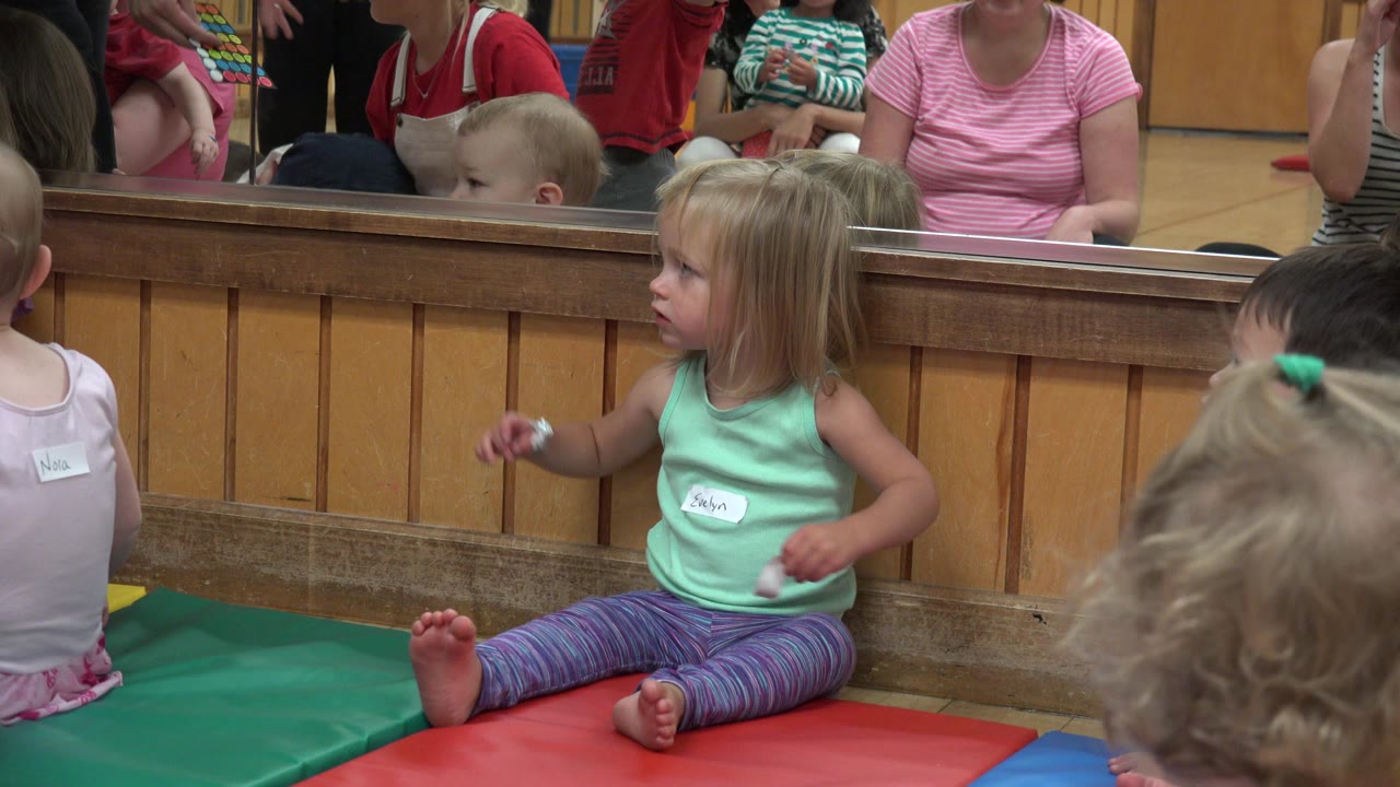 Young blonde girl sitting on colorful floor mats beside other children.