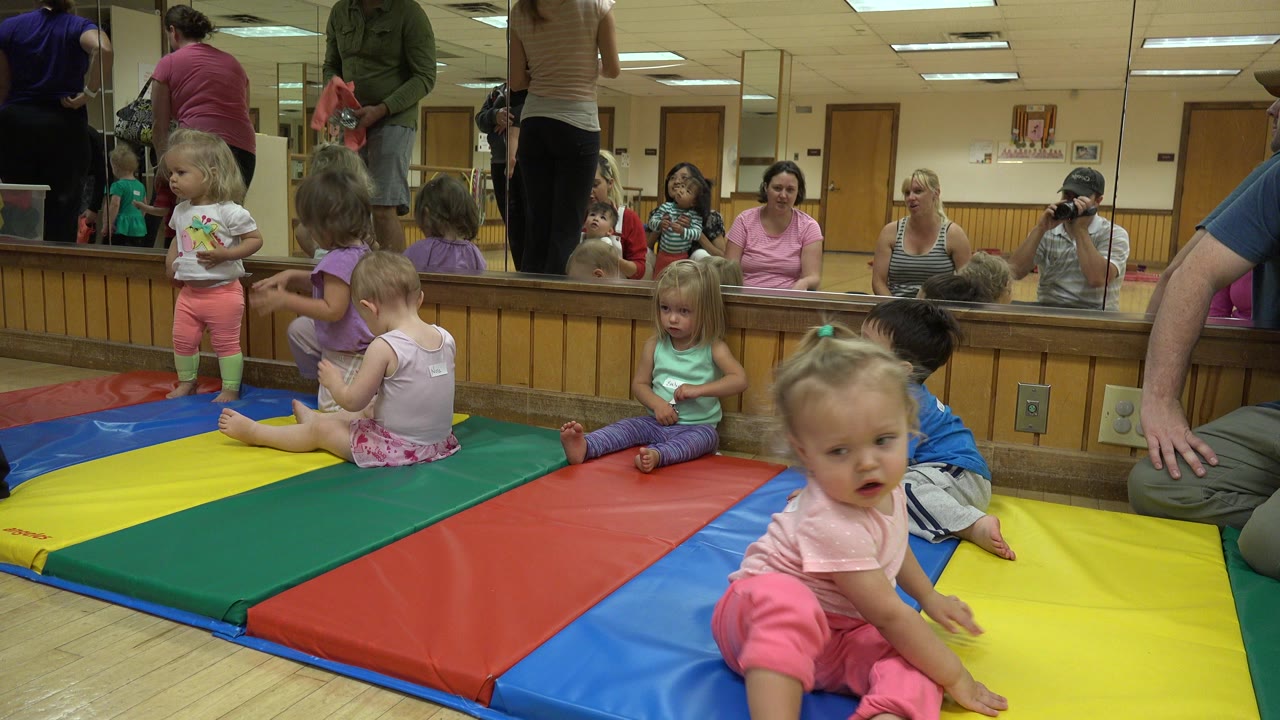 Toddlers sit and stand on red, yellow, green, and blue mats in a Westminster, Colorado indoor play gym while adults appear along the mirrored wall.