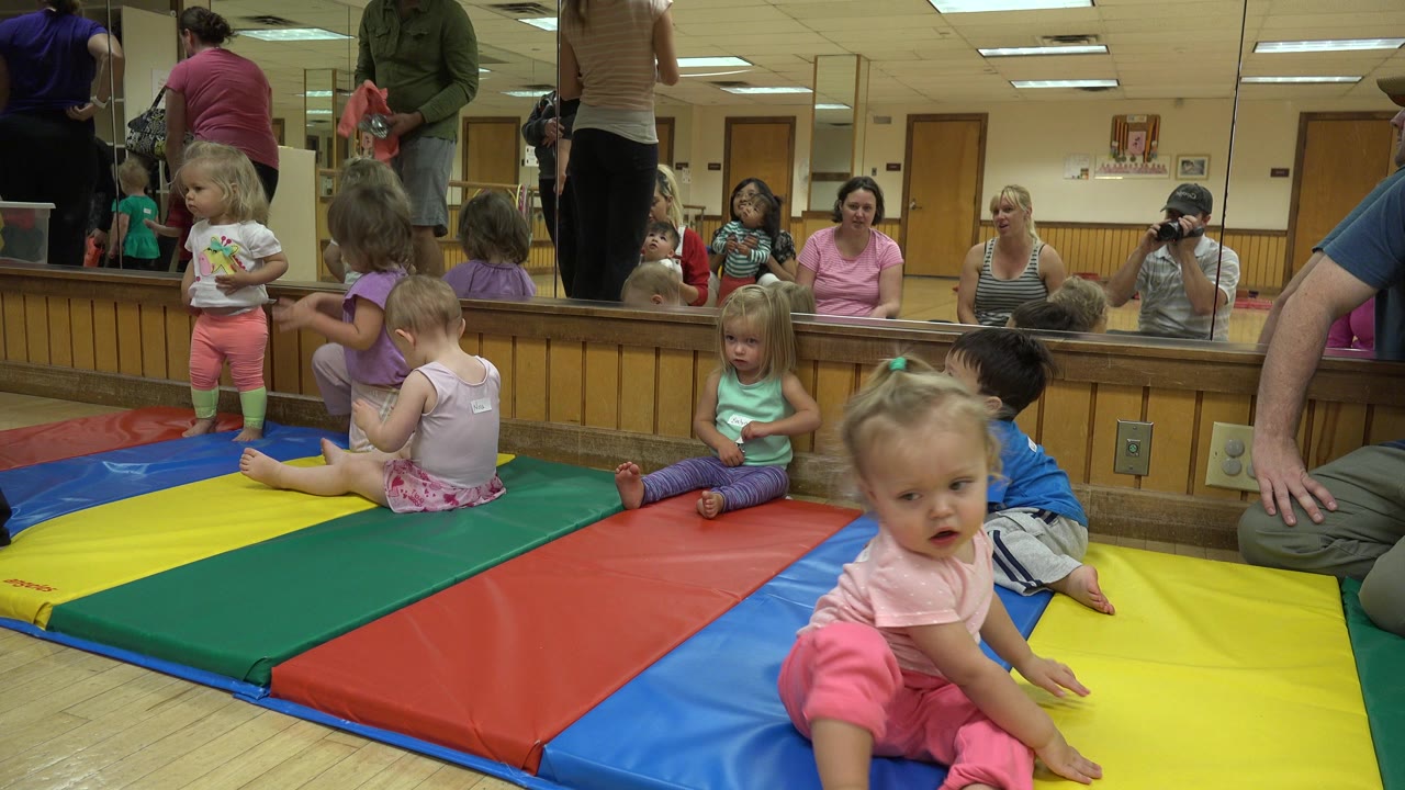Several toddlers remain on the colored mats in the Westminster indoor play space, with seated adults and caregiver reflections visible across the back mirror.