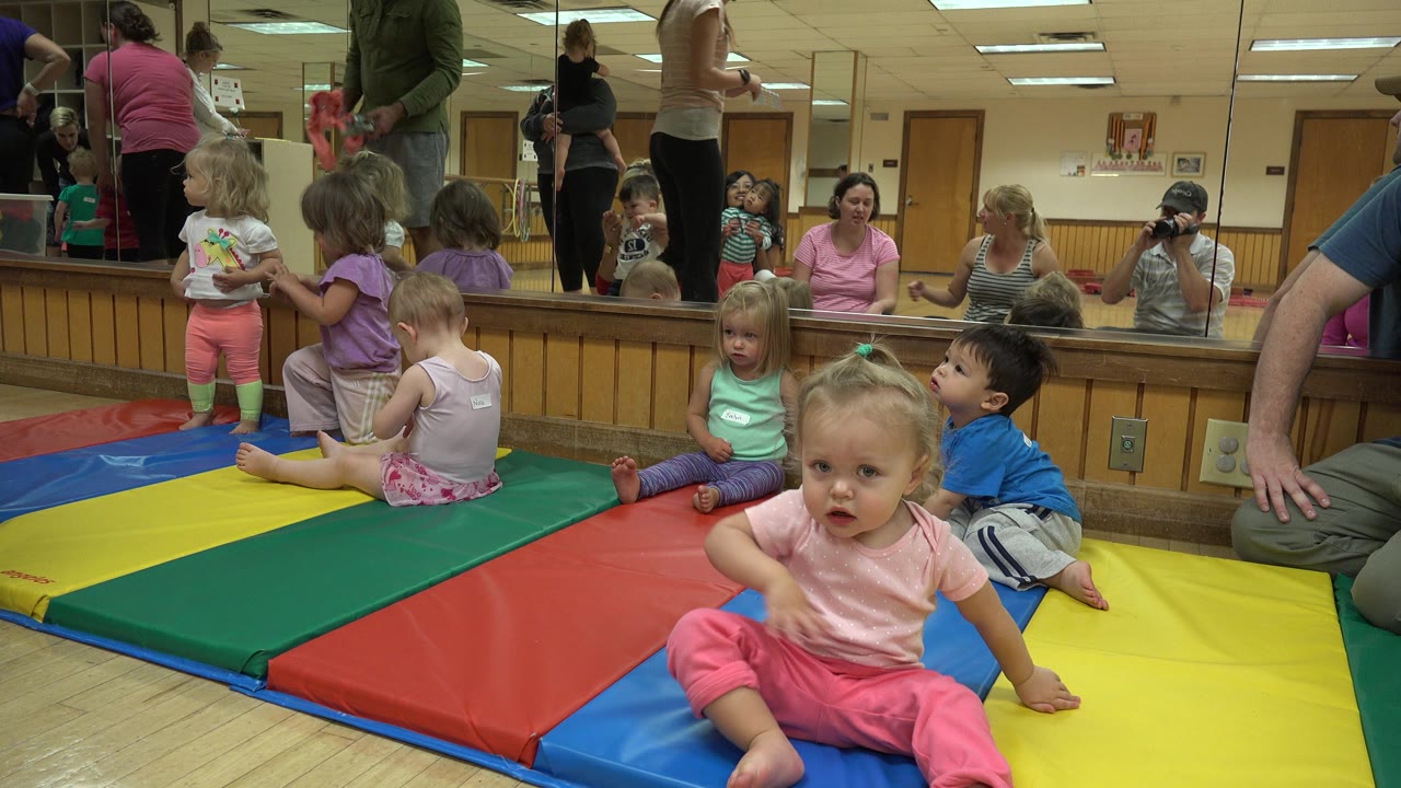 A toddler in pink sits near the foreground while other children play behind her.