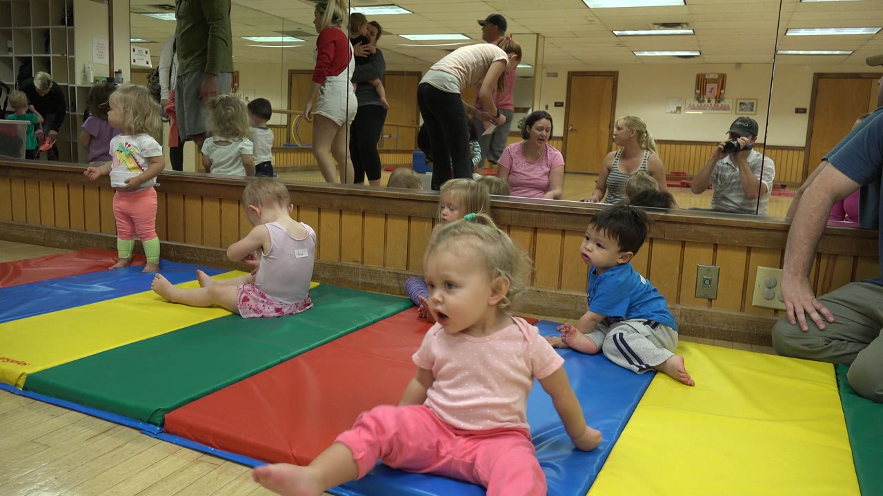 Toddlers on colorful classroom mats with adults reflected in a wall mirror.