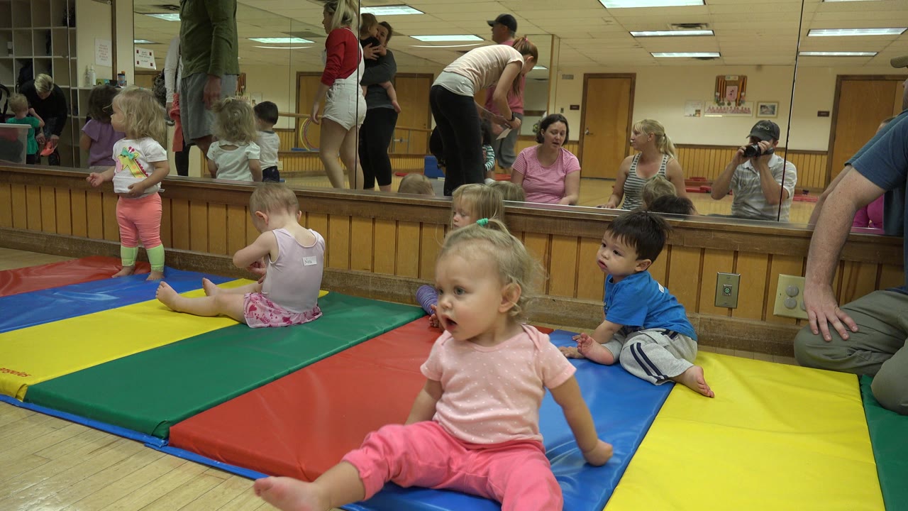A toddler in pink sits near the foreground while other children sit on the mats and adults are reflected across the Westminster classroom mirror.
