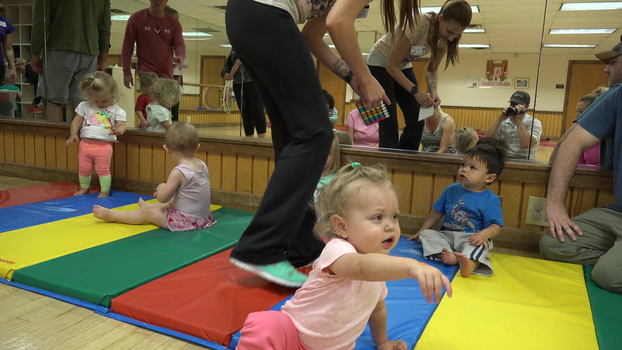 At 0:00 in Westminster, Colorado, toddlers sit and stand on colorful padded mats while adults gather nearby and a mirrored wall reflects the indoor exercise room.