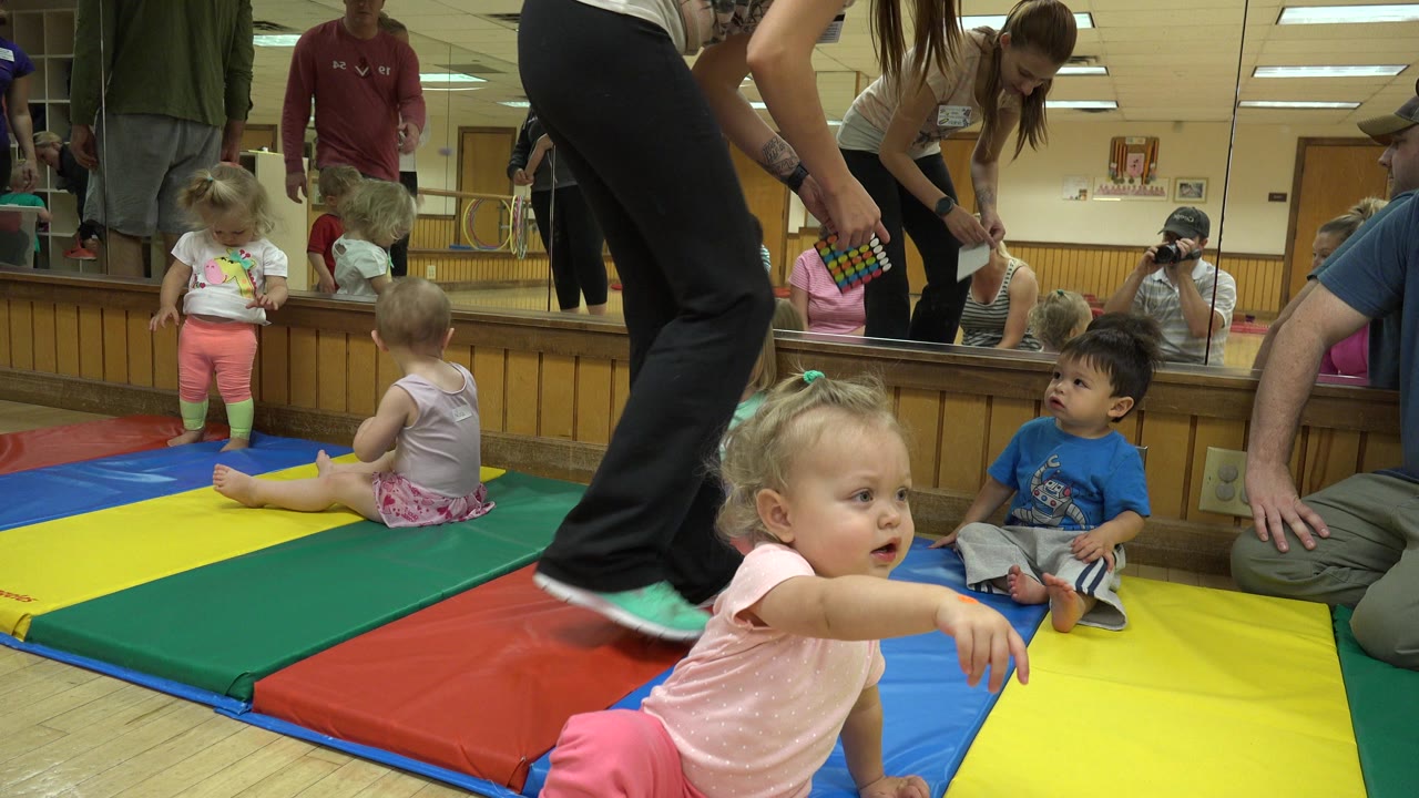 At 0:01 in Westminster, Colorado, a toddler in pink reaches across the foreground on a yellow mat as other toddlers sit near the mirror and adults stand or kneel around them.