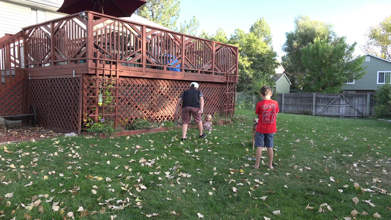 Two children stand on a leaf-scattered lawn in a Westminster, Colorado backyard, with a raised wooden deck and fence behind them.