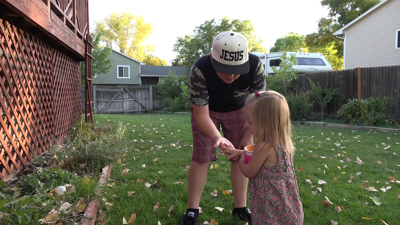 Older child leaning toward a young girl holding cups in a backyard.