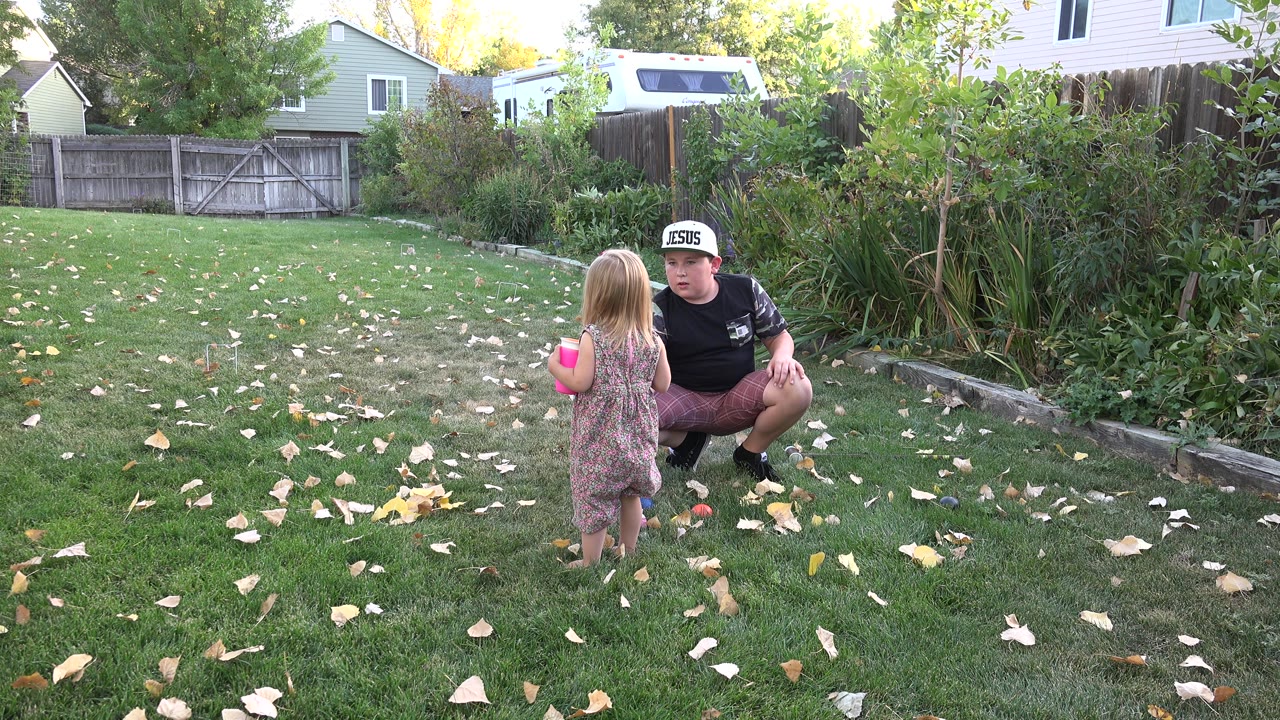 The child holds a bright pink cup while standing on the lawn as the older youth crouches beside colorful toys in the Westminster backyard.