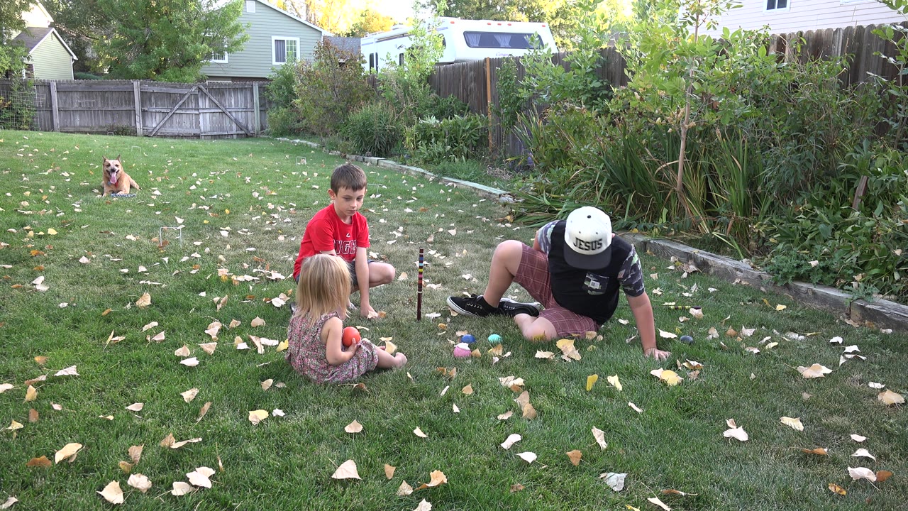 In Westminster, Colorado, a blonde child sits facing small toys on the grass as a boy in a red shirt and an adult in a white hat remain close by in the yard.