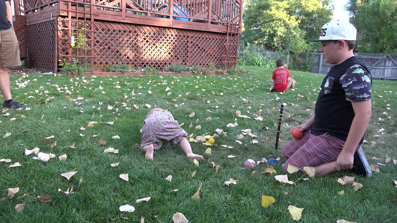In Westminster, Colorado, the young child remains low on the lawn while the older child kneels near small toys, with the deck lattice and backyard fence in the background.