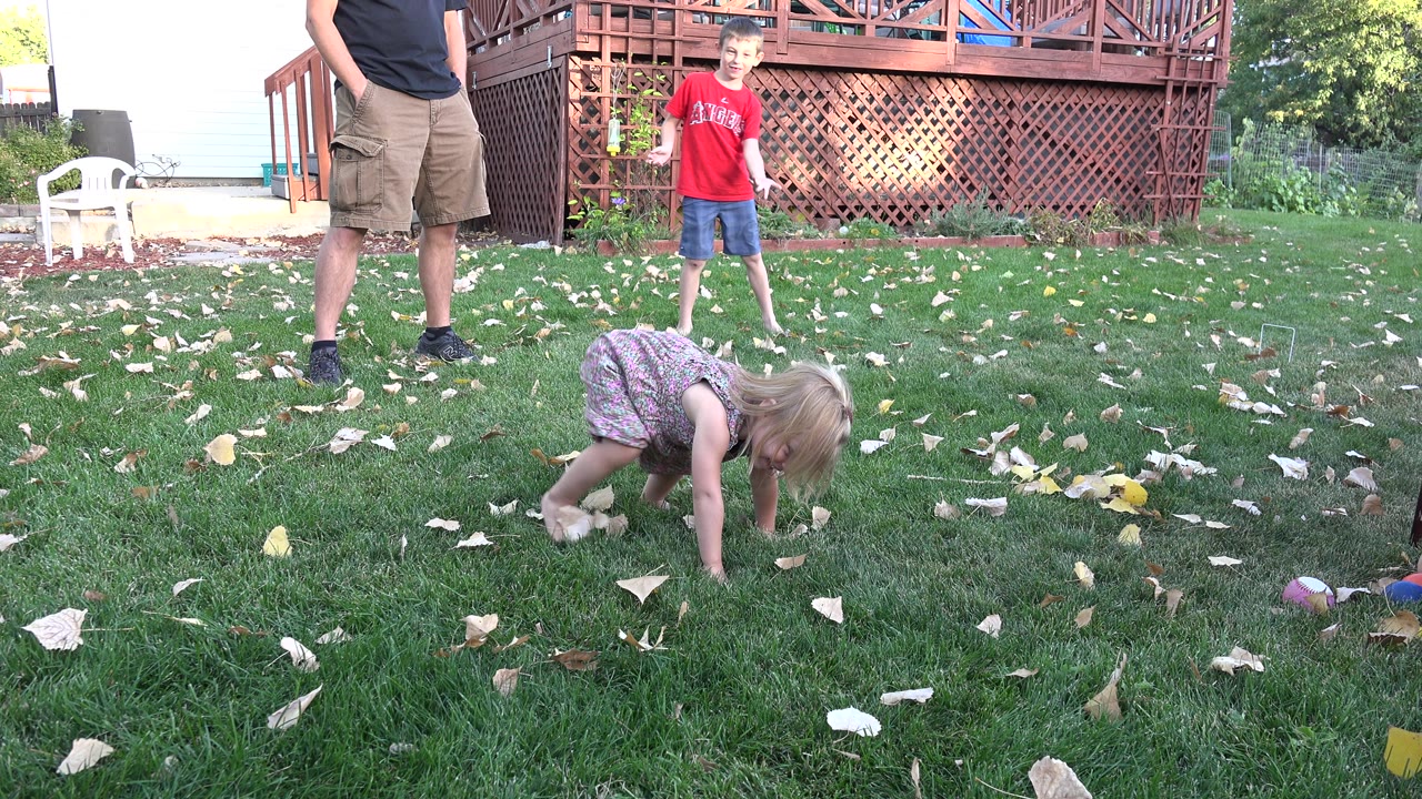 Toddler girl crouches on grass near a boy and adult legs in a backyard.