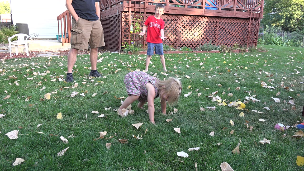 In Westminster, Colorado, the toddler girl bends closer to the grass while the boy in red stands behind her and an adult remains partly cropped at left.