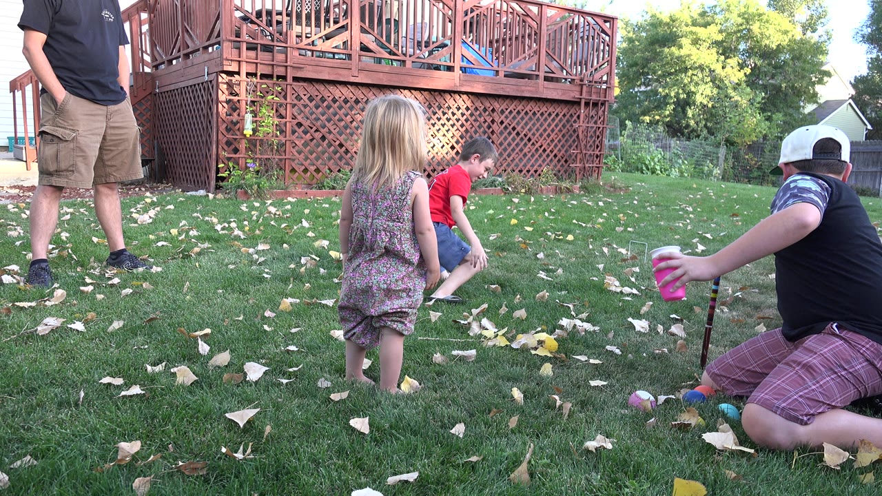 In Westminster, Colorado, children gather on a grassy backyard lawn while a partially visible adult stands near a red-brown wooden deck.