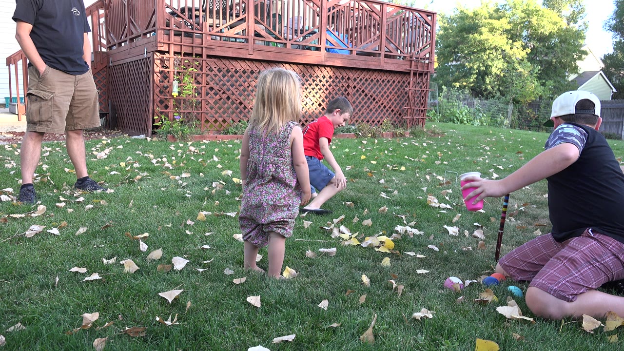 In Westminster, Colorado, a young girl stands with her back to camera as two boys play on the lawn near scattered toys under a raised deck.