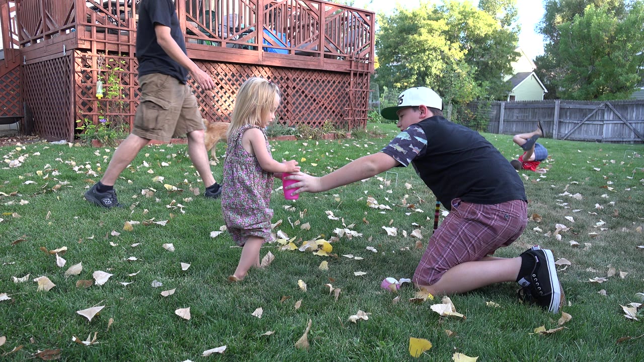 In Westminster, Colorado, the girl holds a pink cup while the kneeling boy reaches toward her across the grass, with a deck, fence, leaves, and backyard toys behind them.
