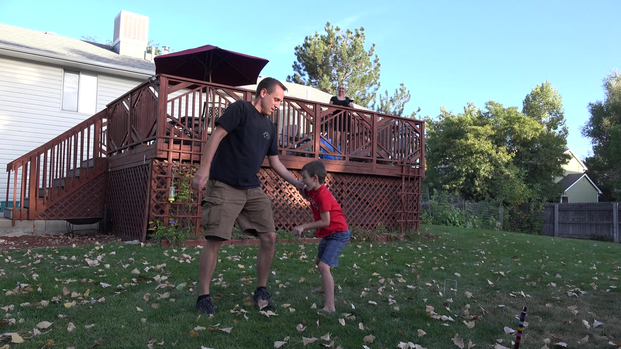 An adult man leans toward a young boy in a red shirt on the Westminster backyard lawn, with the raised deck and house behind them.