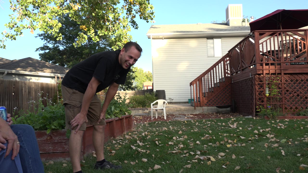 A younger adult man leans forward in a Westminster, Colorado backyard while seated adults are partly visible at the frame edges near grass, garden beds, a deck, and a house.