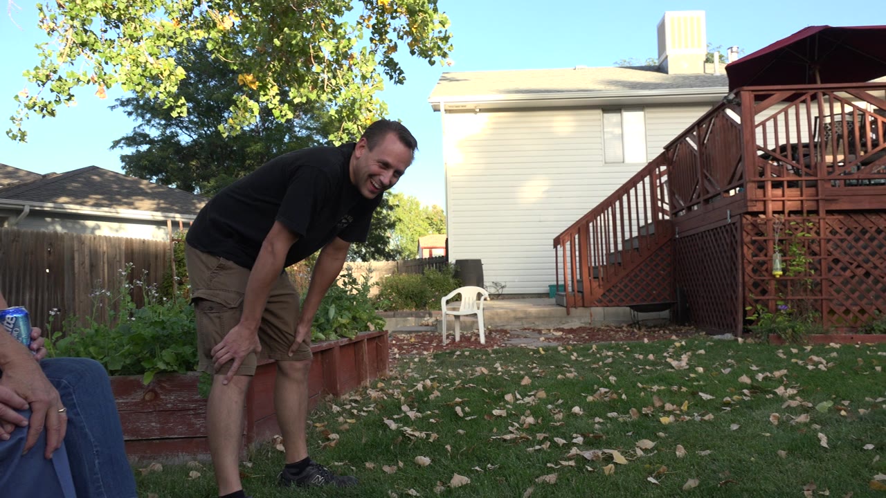 The younger adult man remains bent forward in the Westminster backyard, with a raised garden bed, wood deck, fence, trees, and a light-colored house behind him.