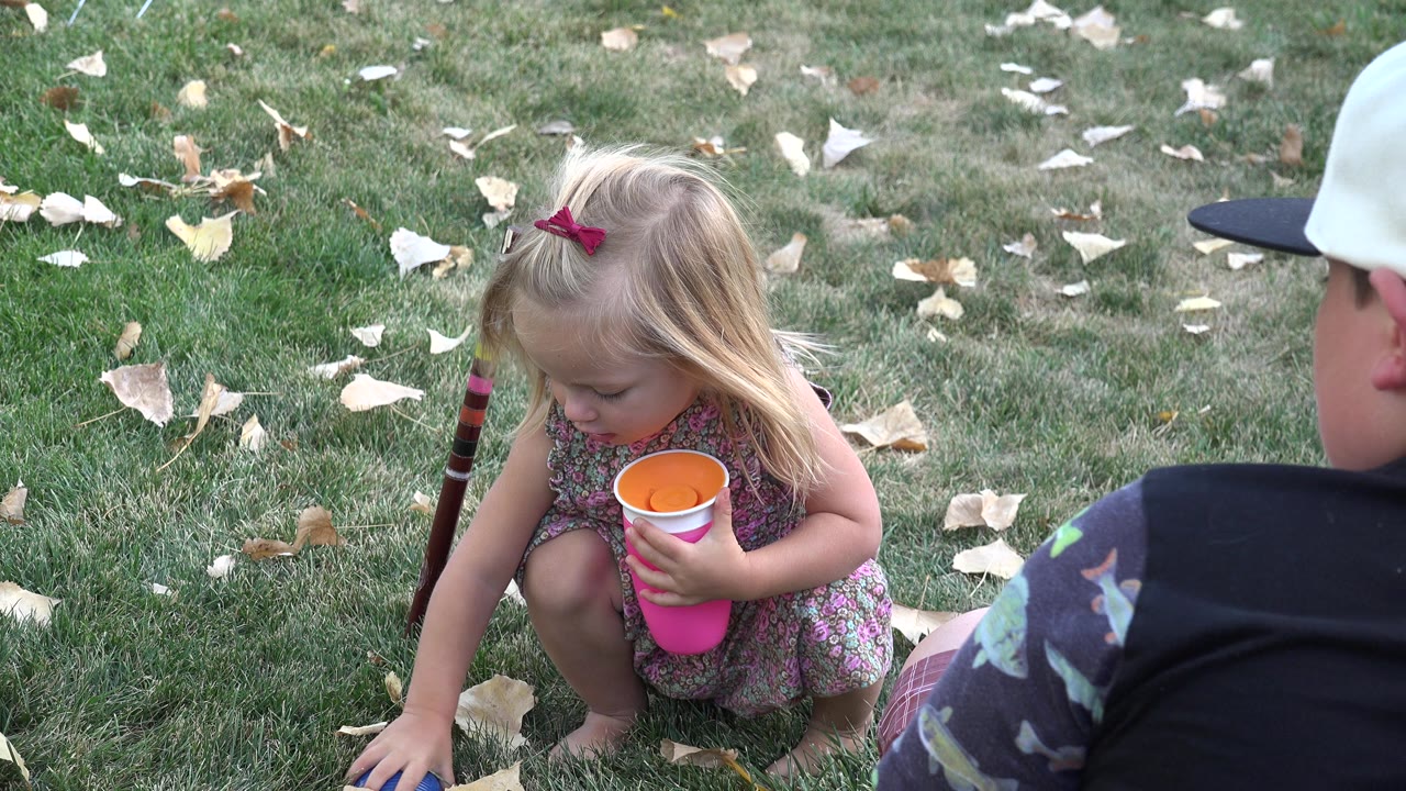 Young girl crouches on grass holding a pink cup near a seated boy in a white cap.