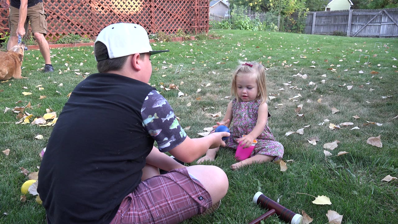 Boy in a white cap hands a blue toy to a small girl sitting on grass.