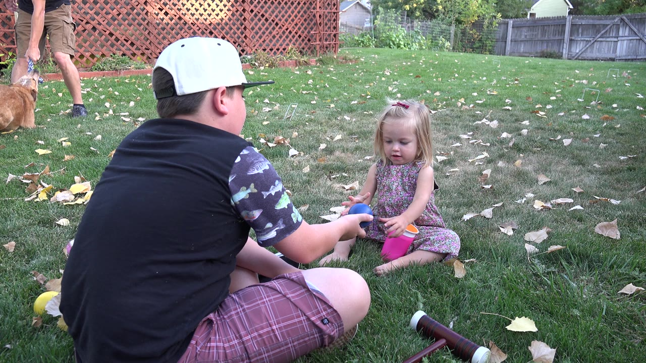 The Westminster backyard scene holds on the boy seated in the foreground and the girl across from him, with scattered leaves, toys, fence panels, and a partial adult with a dog behind them.