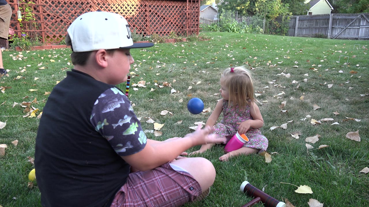 Boy in a white cap sits on grass facing a little girl with toys nearby.