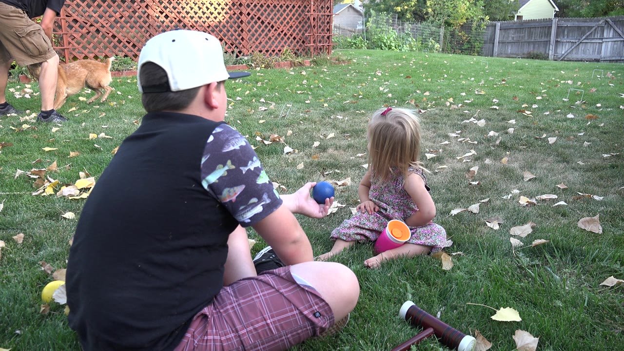 Boy in a white cap sits on grass near a little girl during backyard croquet play.