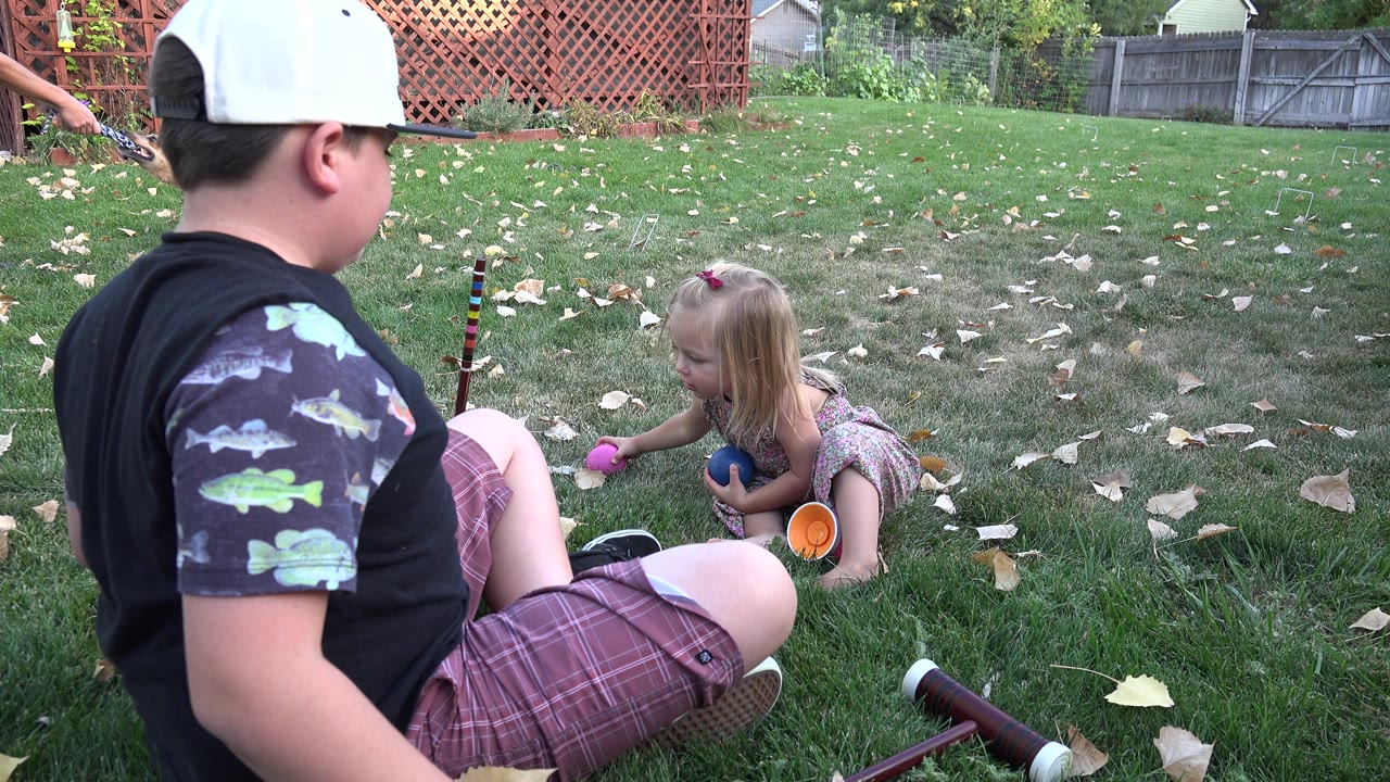 Seated older child or adult facing a young girl on a backyard lawn with toy balls nearby.