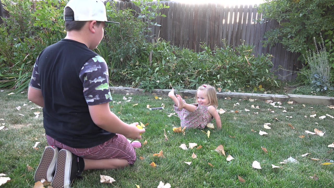 Kneeling person faces a young girl seated on grass in a backyard.