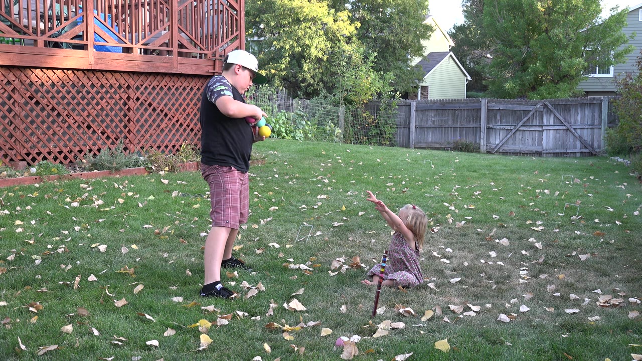 Older boy or young man holding colorful balls near a seated young girl in a backyard.