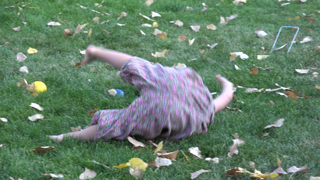 Young girl rolling sideways on green grass with fallen leaves.