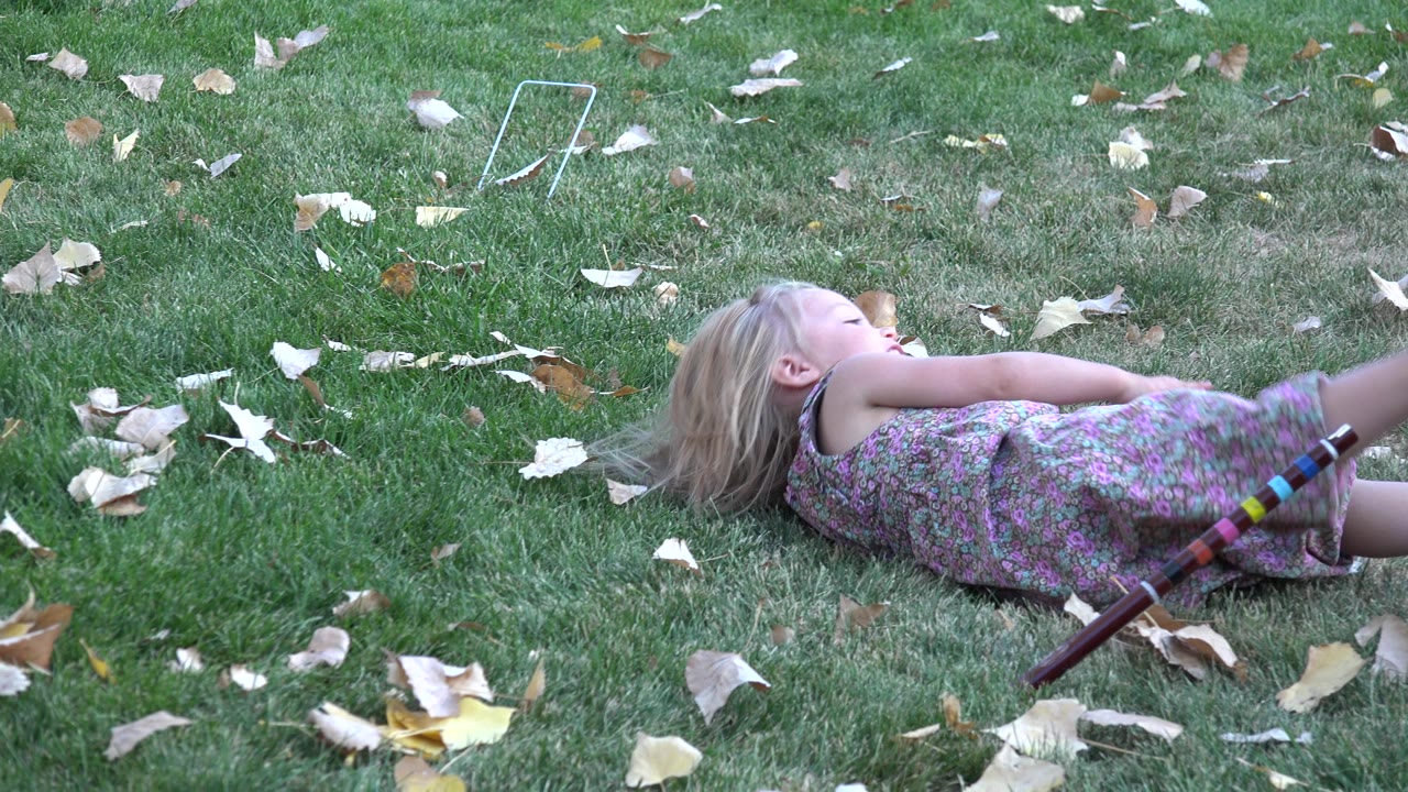 Young blonde girl in a floral dress lies on a leaf-strewn lawn in Westminster, Colorado, with a striped stick or toy resting near her legs.