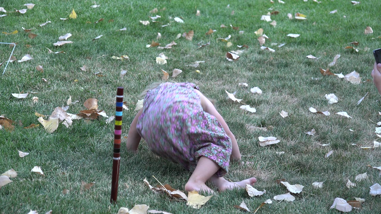 Young girl bent forward on grass near fallen leaves.