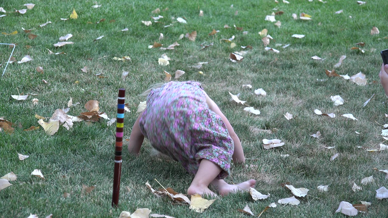 The child remains crouched on the grassy Westminster scene, with one leg extended behind her, pale leaves on the lawn, and the striped pole near the center foreground.