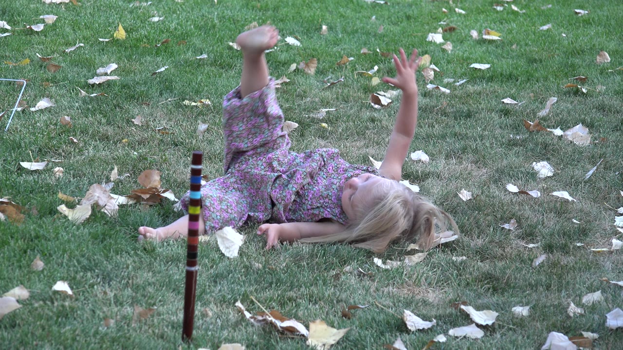 A young blonde child lies on a green lawn in Westminster, Colorado, with one leg raised and a striped yard stake standing in the foreground.
