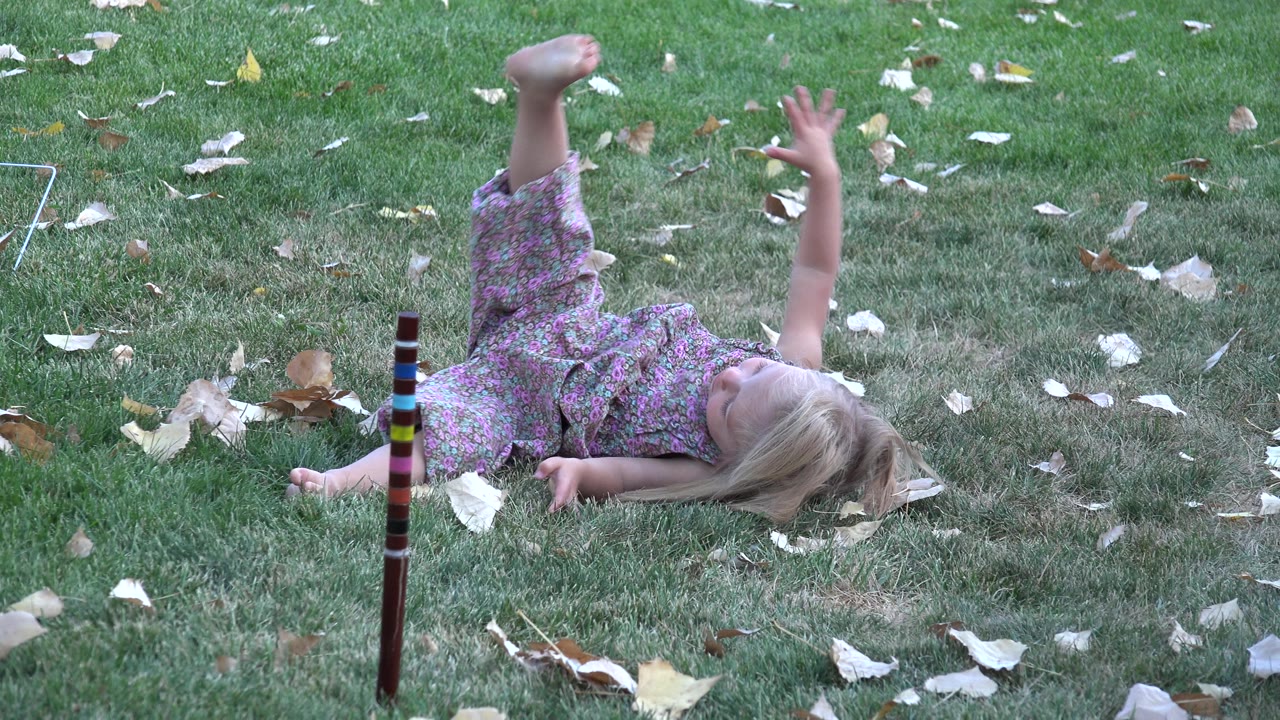 The child remains on their back on the Westminster lawn, one hand lifted in the air while dry leaves dot the surrounding grass.