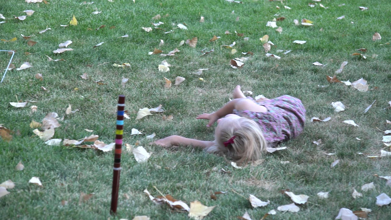 A small blonde girl in a patterned dress lies sideways on a leaf-strewn lawn in Westminster, Colorado, with a striped stake in the foreground.