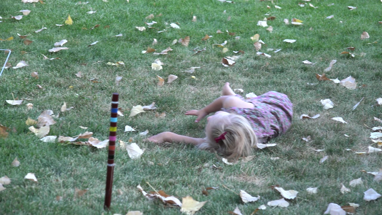 The blonde child continues rolling across the grass in Westminster, Colorado, with her head lowered and dry leaves spread around her.