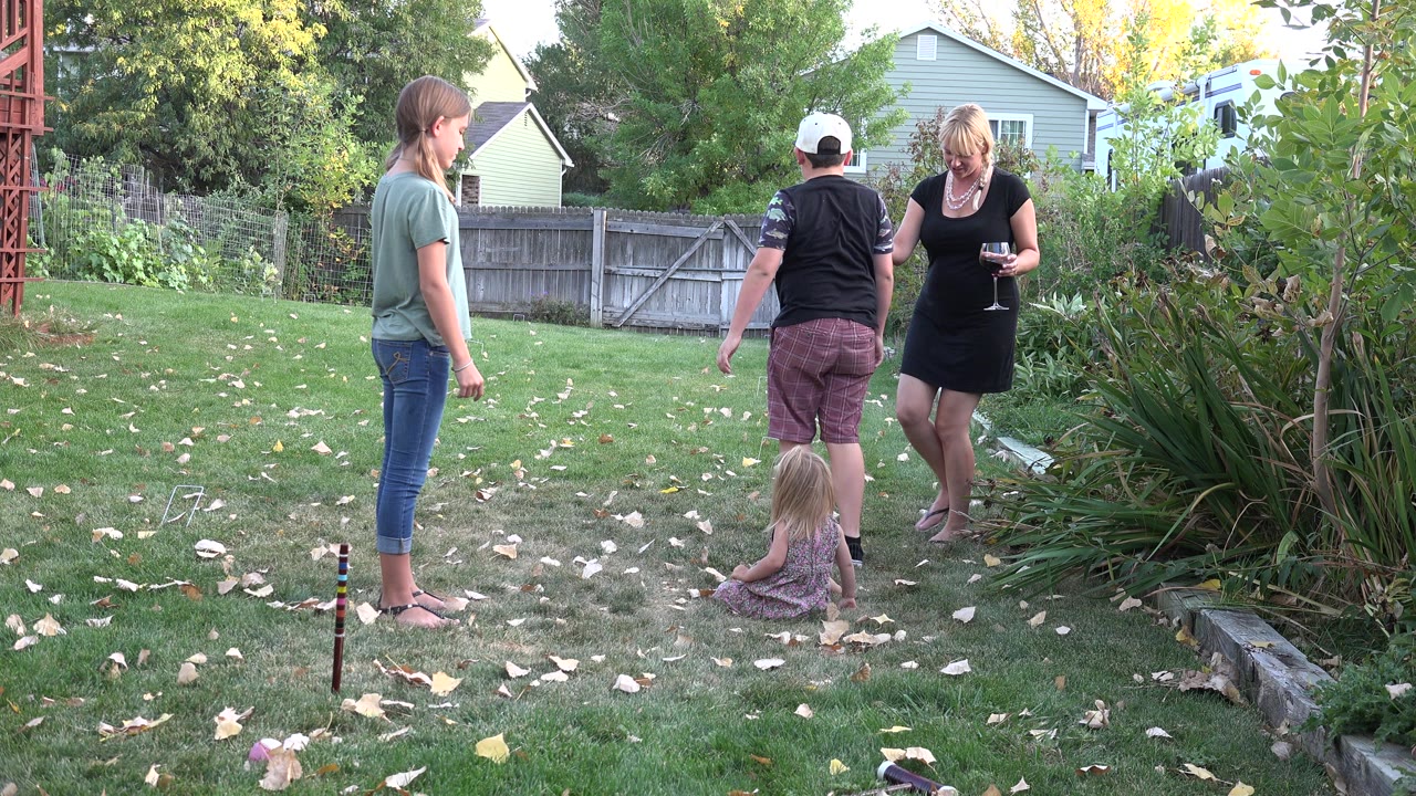 Family members in a grassy backyard with a seated child and garden plants.