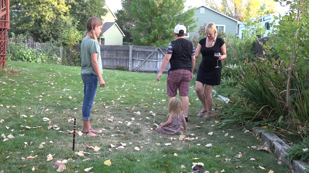 In Westminster, Colorado, the group stays in the backyard as the seated child faces the adults and a woman in a black dress stands near the garden bed holding a glass.