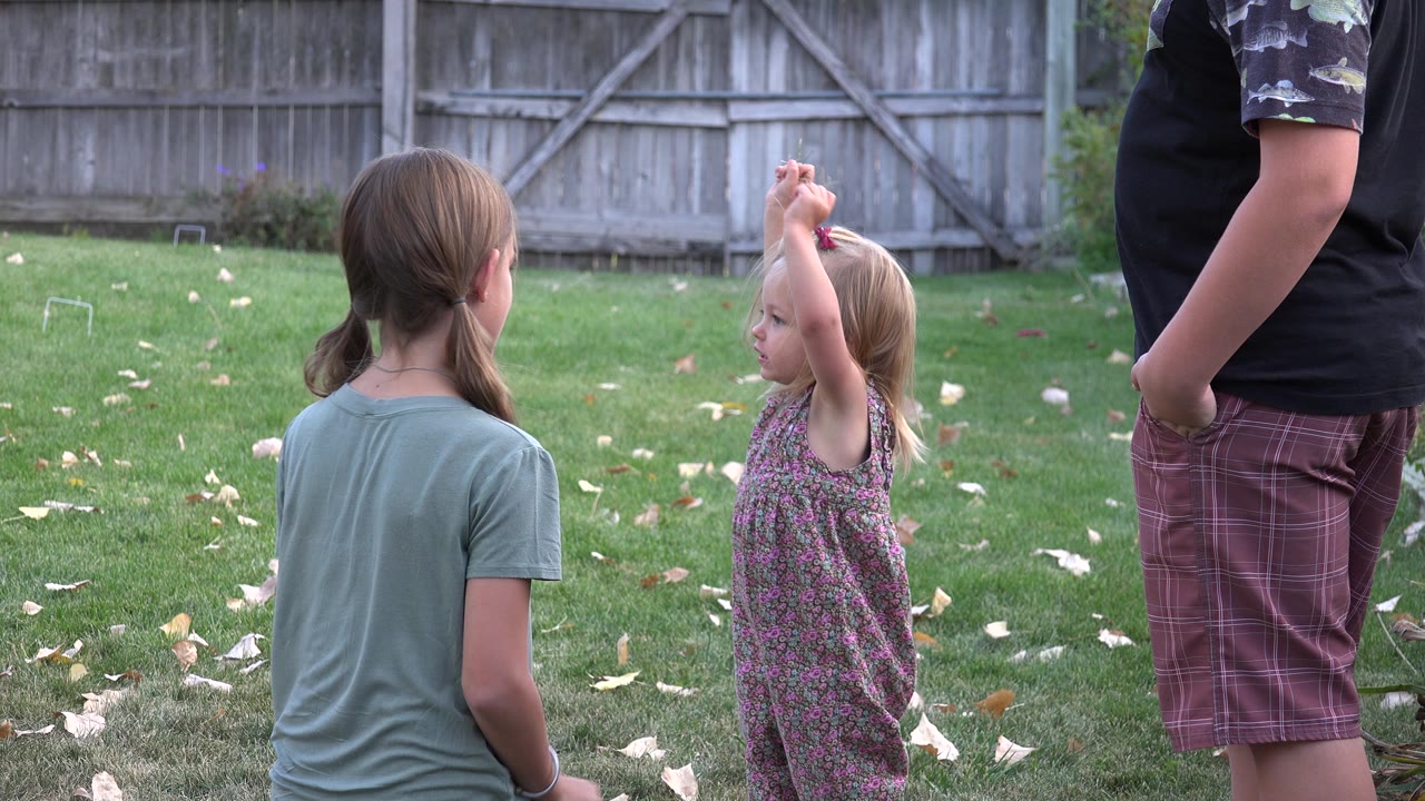 In Westminster, Colorado, the smaller girl lifts both arms overhead as an older girl sits or crouches nearby and the adult remains partly cropped at frame right.