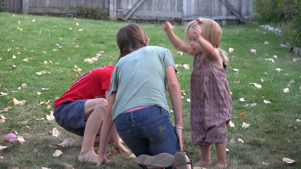 Three children playing on a backyard lawn near a wooden fence.