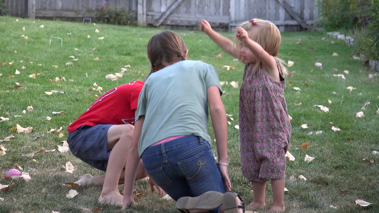 In the Westminster backyard, two older children crouch near the grass while the younger girl in a patterned dress raises both arms beside them.