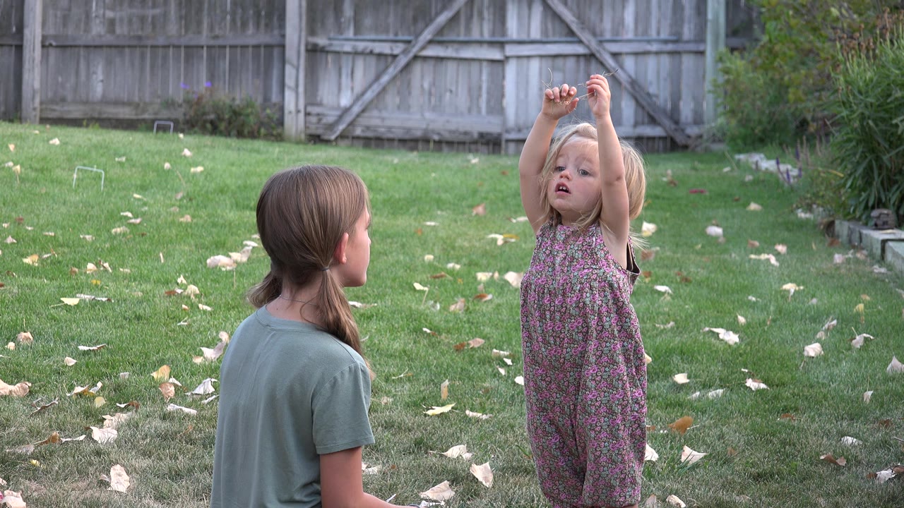 Two children on a grassy lawn, one seated or crouched and one raising her arms.