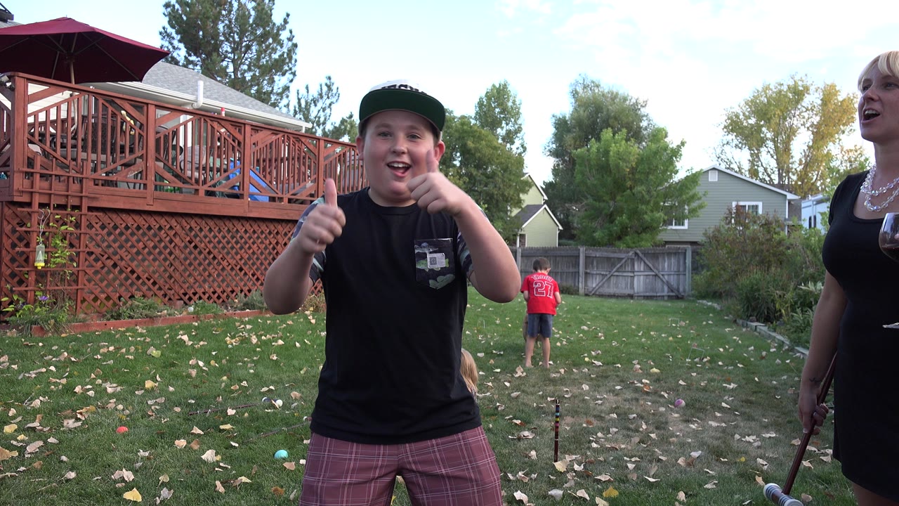 Boy gives two thumbs up in a backyard near a woman holding a mallet.