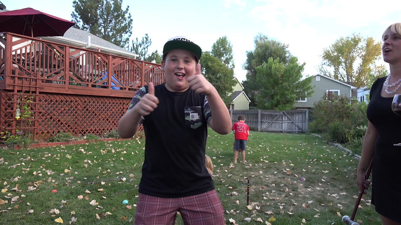 The boy remains near the camera giving thumbs up in the Westminster backyard, with the woman in a black dress partly visible and another child farther back on the lawn.