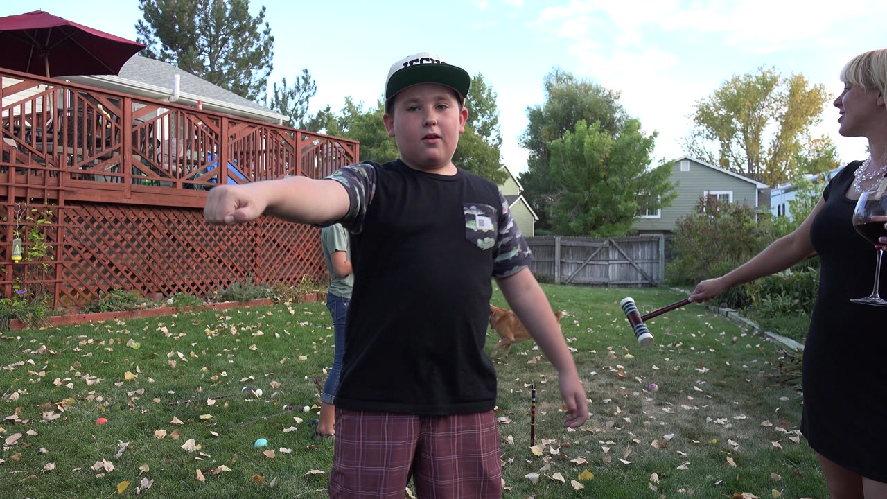Boy in a cap stands on a backyard lawn near an adult holding a mallet.