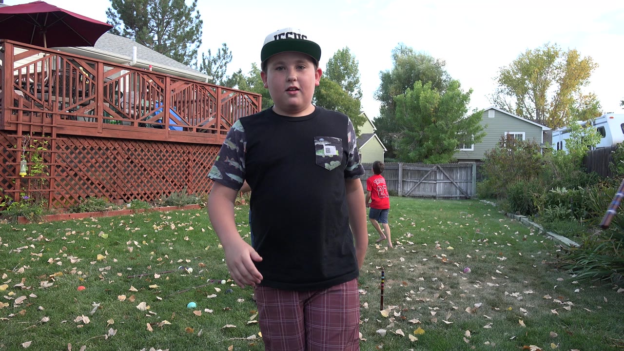 The boy remains centered in the Westminster backyard as another child in a red shirt appears in the background near the fence and shrubs.