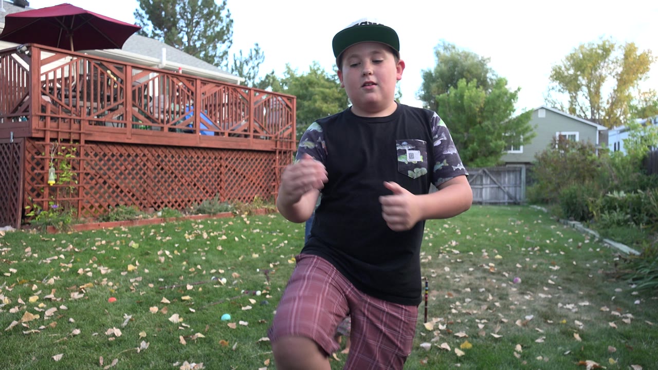 The boy raises one hand while facing forward in the Westminster backyard, with scattered leaves on the grass and the red deck railing filling the left background.